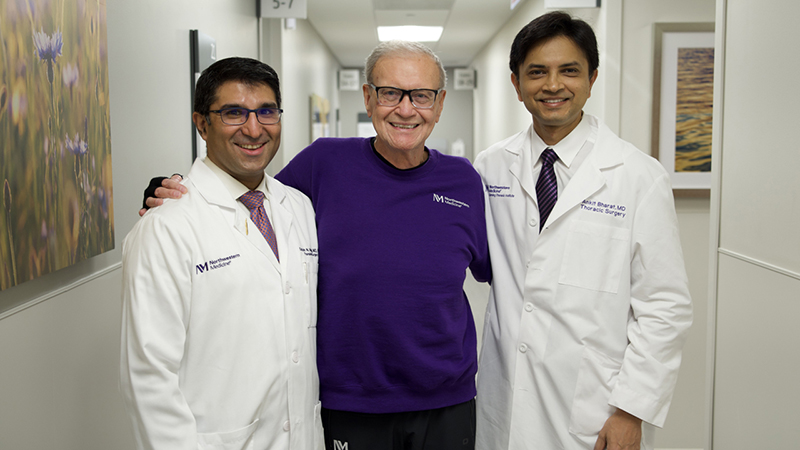 Posing arm in arm in a hospital corridor, from left to right: Satish Nadig, MD, PhD, wearing a white lab coat; patient Gary Gibbon, MD in a purple sweatshirt; and Ankit Bharat, MD, in a white lab coat.