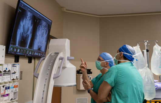 Two Northwestern Medicine orthopaedic surgeons wearing green scrubs in an operating room pointing to an X-ray of a hip on a monitor.