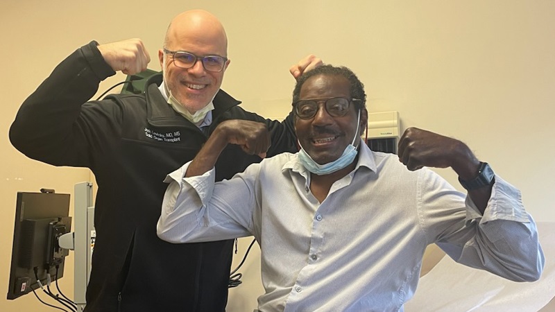A black male double organ transplant recipient sitting on a hospital bed while flexing his arm muscles next to a white male physician wearing a black jacket.