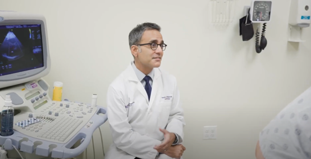 Dr. Sanjiv Shah speaking with a patient while sitting next to an ultrasound machine in an exam room 
