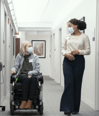Black woman in a long skirt walking down the hall, talking to a blonde white woman in a wheelchair.