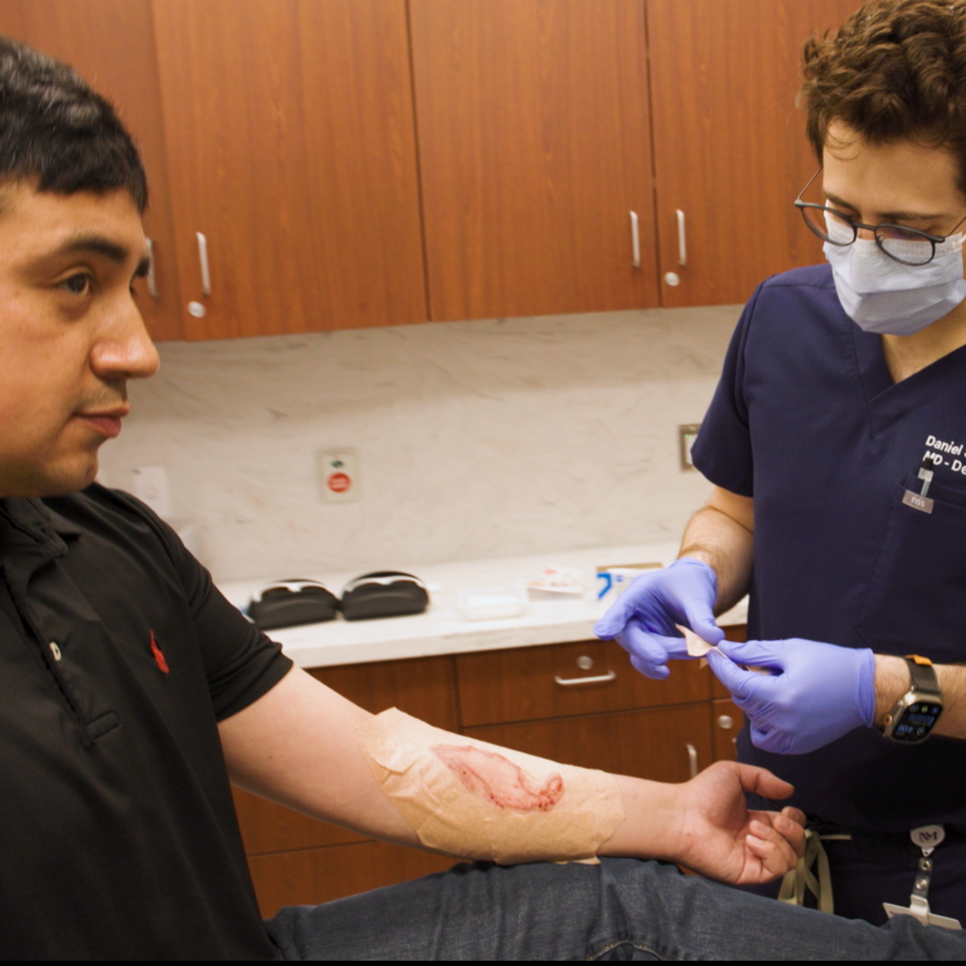 Patient in dermatology office being treated for a skin condition. The patient has his arm out, and a doctor and another clinician are treating him.