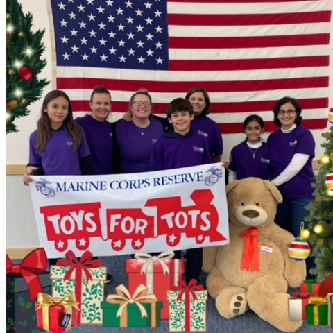 A group of women and a young boy and girl standing behind Christmas presents and in front of an American flag while holding a sign that says Toys For Tots.