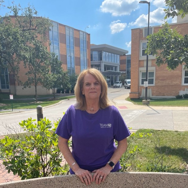 Woman in purple NM shirt standing in a courtyard. She is standing between two flower beds.