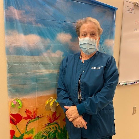 NM lab technician Laura Savant standing in front of a tapestry showing sky and flowers.