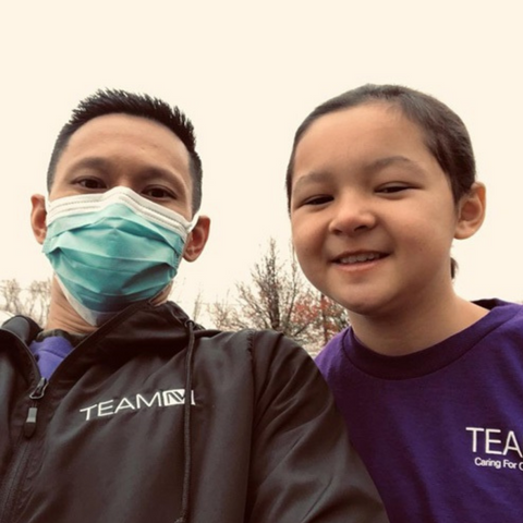 A man with brown hair, wearing a black jacket and medical mask standing next to a young girl wearing a purple shirt and smiling.