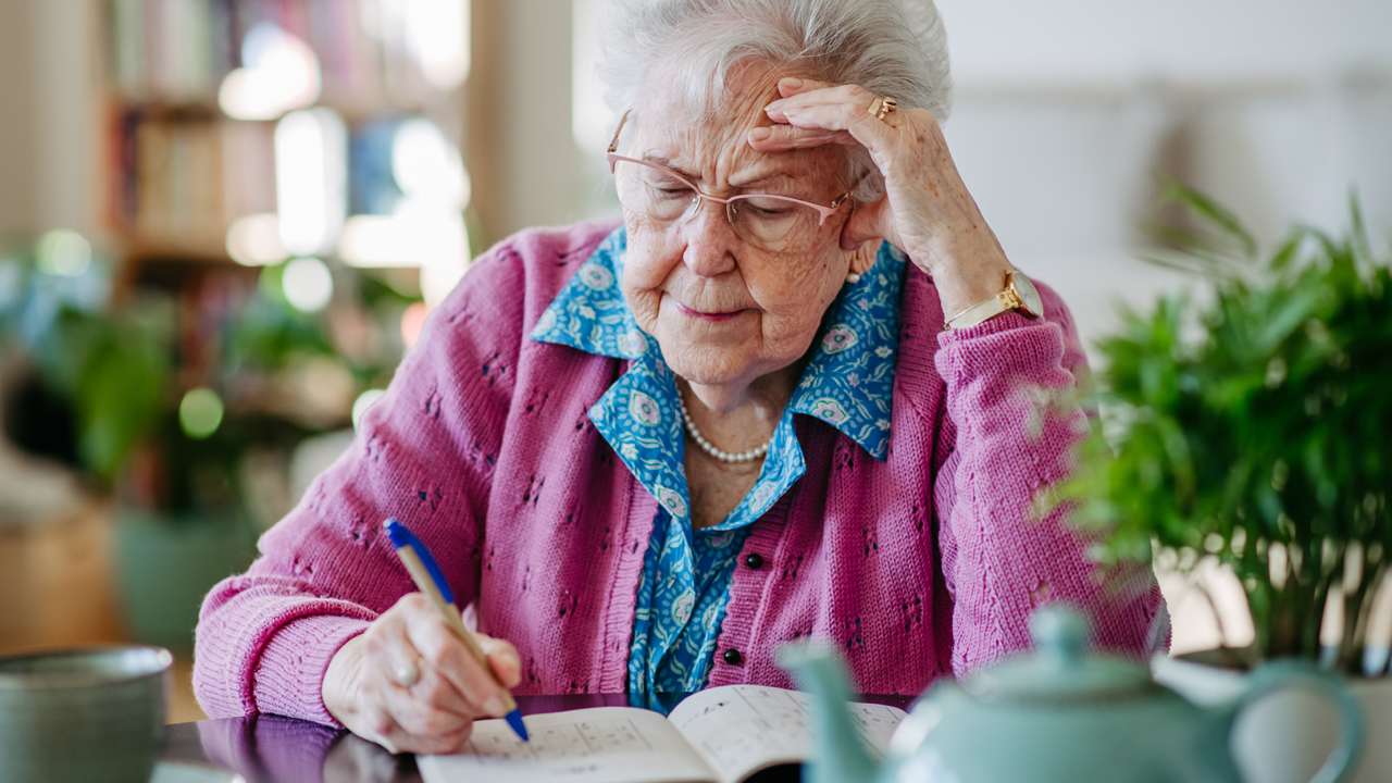 A person with gray hair wearing glasses and a pink sweater holds a pen and solves a sudoku puzzle at a kitchen table. 
