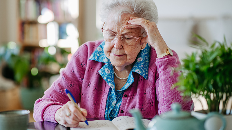A person with gray hair wearing glasses and a pink sweater holds a pen and solves a sudoku puzzle at a kitchen table. 