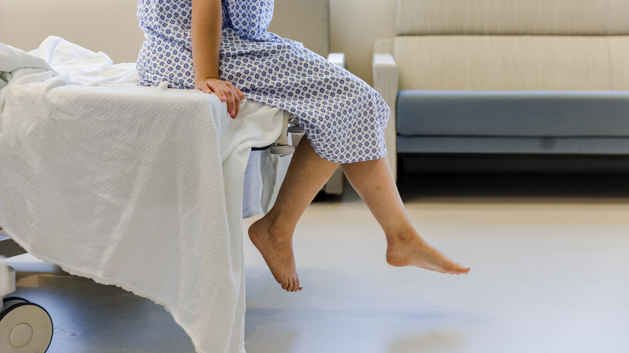 Side profile of a person wearing a hospital gown pictured from the waist down and sitting on the edge of a hospital bed. 