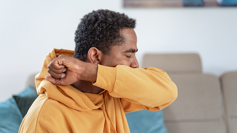 Adult wearing a mustard yellow sweatshirt sitting on a couch and coughing from croup into elbow