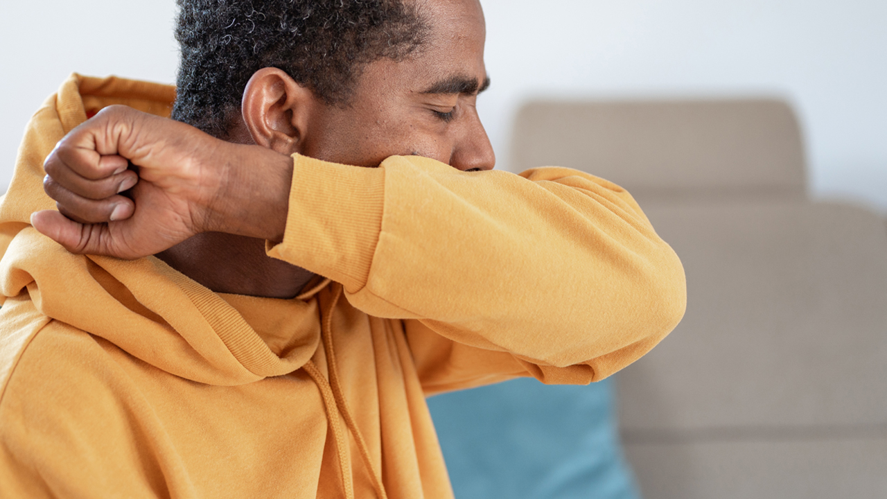 Adult wearing a mustard yellow sweatshirt sitting on a couch and coughing from croup into elbow