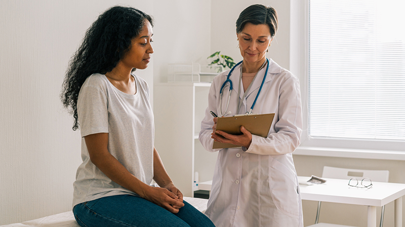 A patient sitting on a table in a medical room, speaking to a physician who is writing notes on a pad and wearing a stethoscope around their neck with a white coat. 