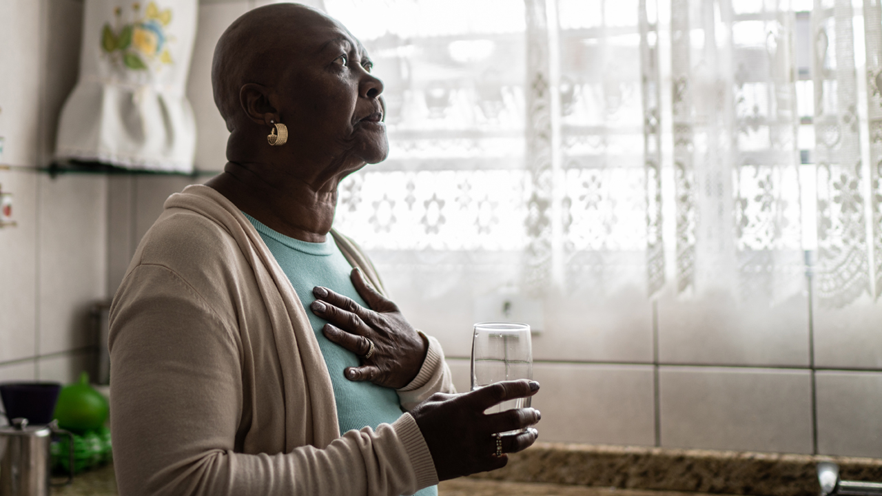 Older adult in a kitchen holding their chest in one hand and a glass of water in the other.