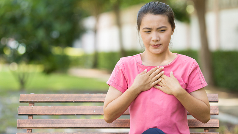 Person wearing a pink shirt, sitting on a park bench holding their chest. 
