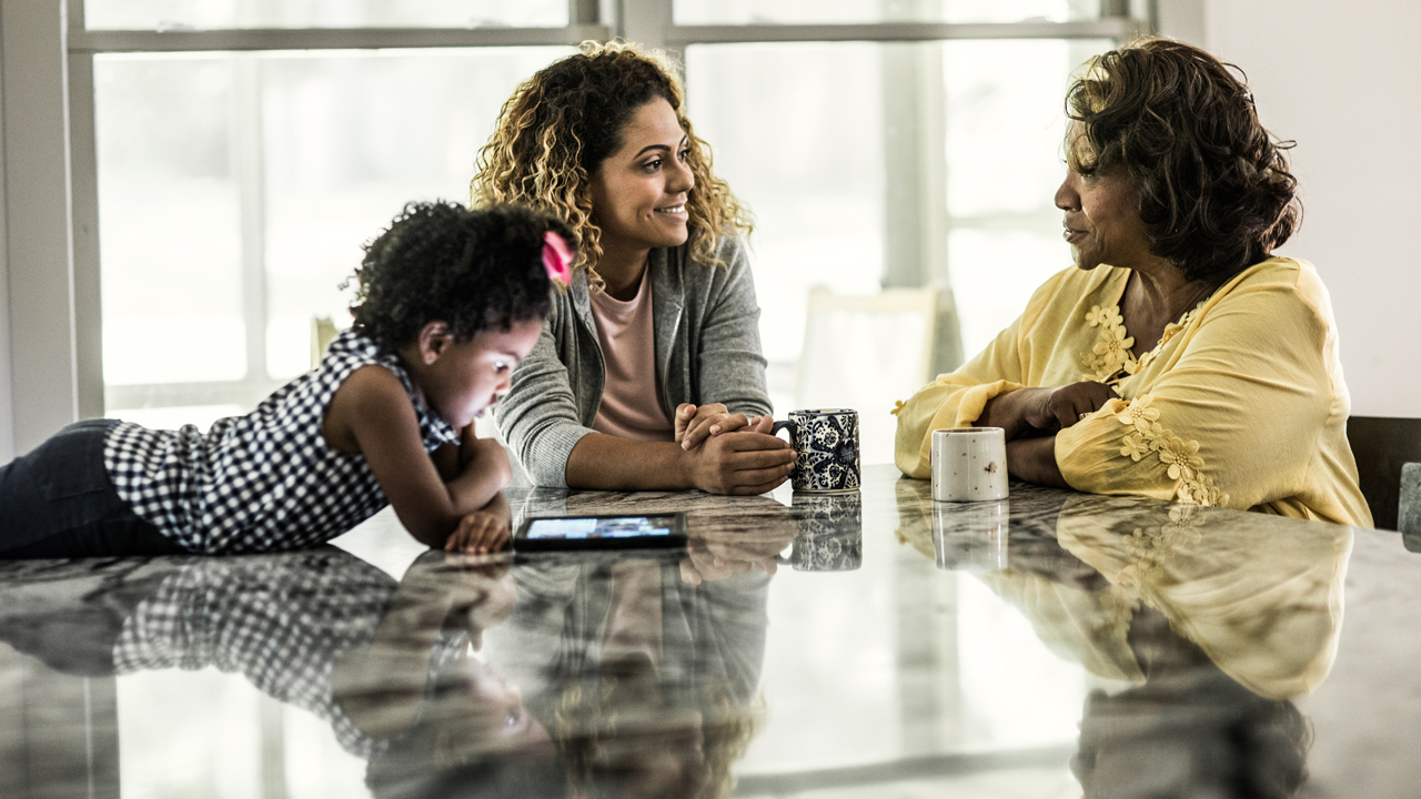 Woman with curly dark hair and medium tan skin smiles at older woman over coffee at a table, while child with black curly hair looks at an iPad.