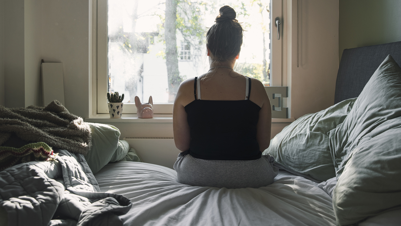Woman sitting on unmade bed, peering out the window, viewed from the back.