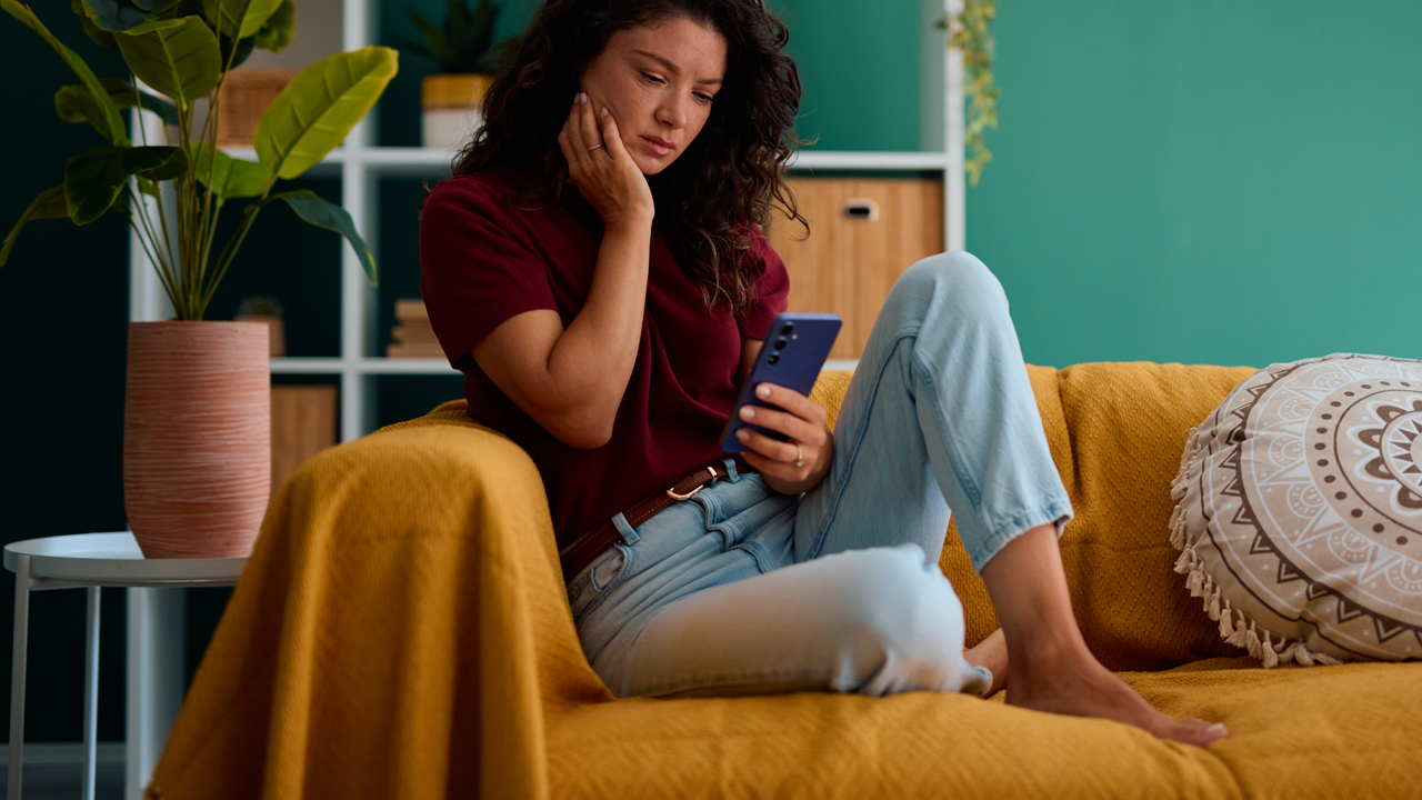 A person with long, curly black hair looks worried while looking at a smartphone and sitting on a couch. 