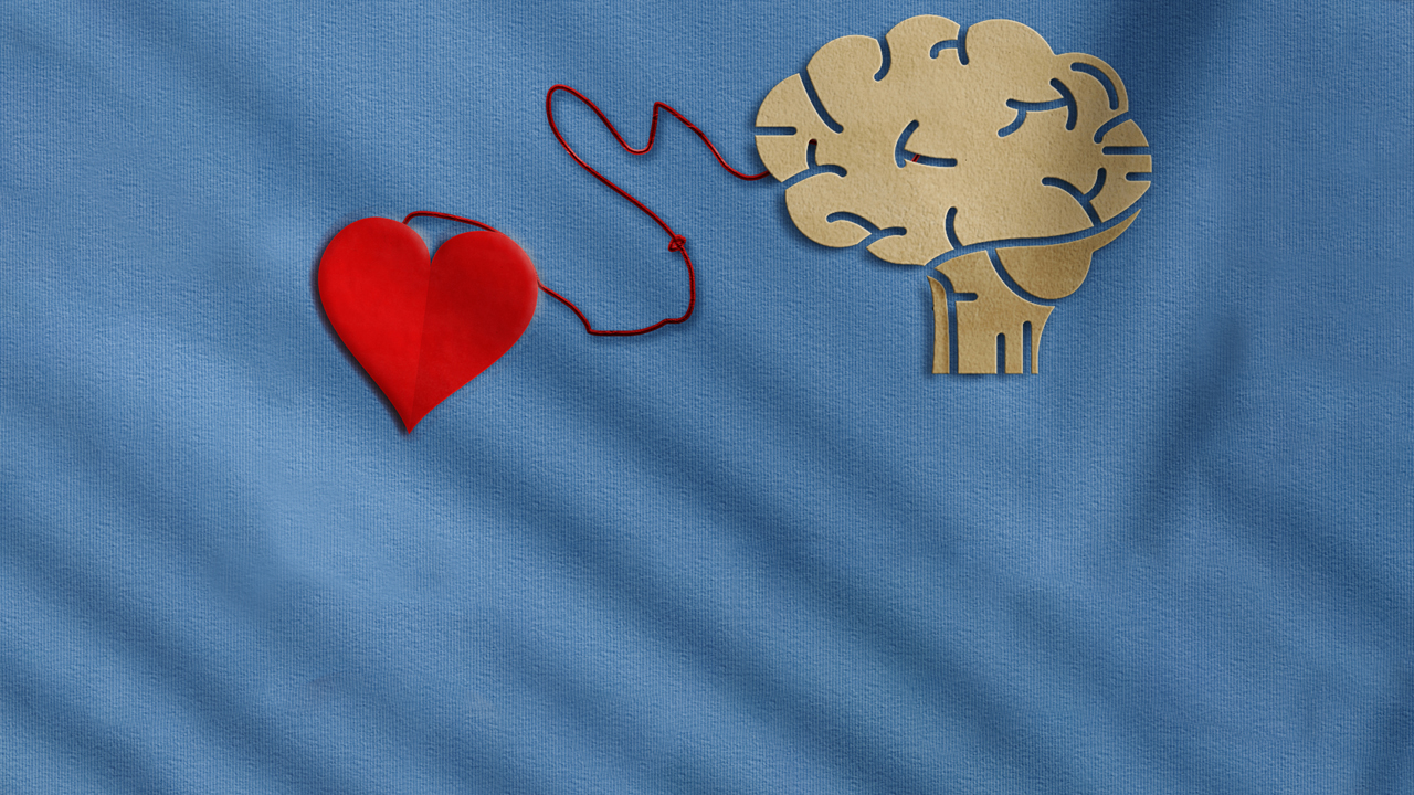 Overhead image of a heart shape, made out of fabric, attached by a red string to a cardboard cutout of a brain all over a light blue fabric background.