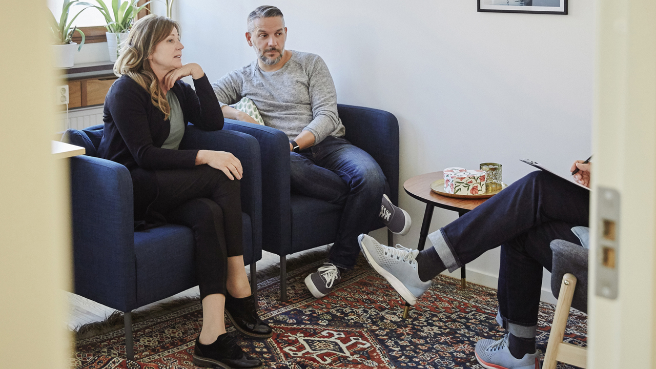 A couple sits in two blue chairs facing a therapist writing on a clipboard.