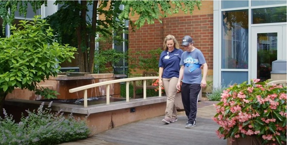 A patient in a blue shirt and baseball cap walking with an occupational therapist over a short bridge.