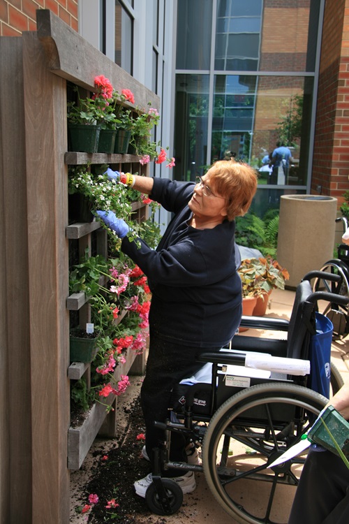 Patient with red hair planting flowers while standing next to her wheelchair.