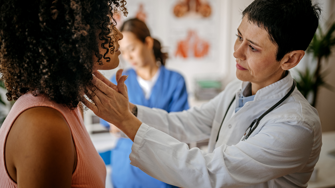 A physician examining a patient's thyroid with fingers on both sides of the patient's neck,