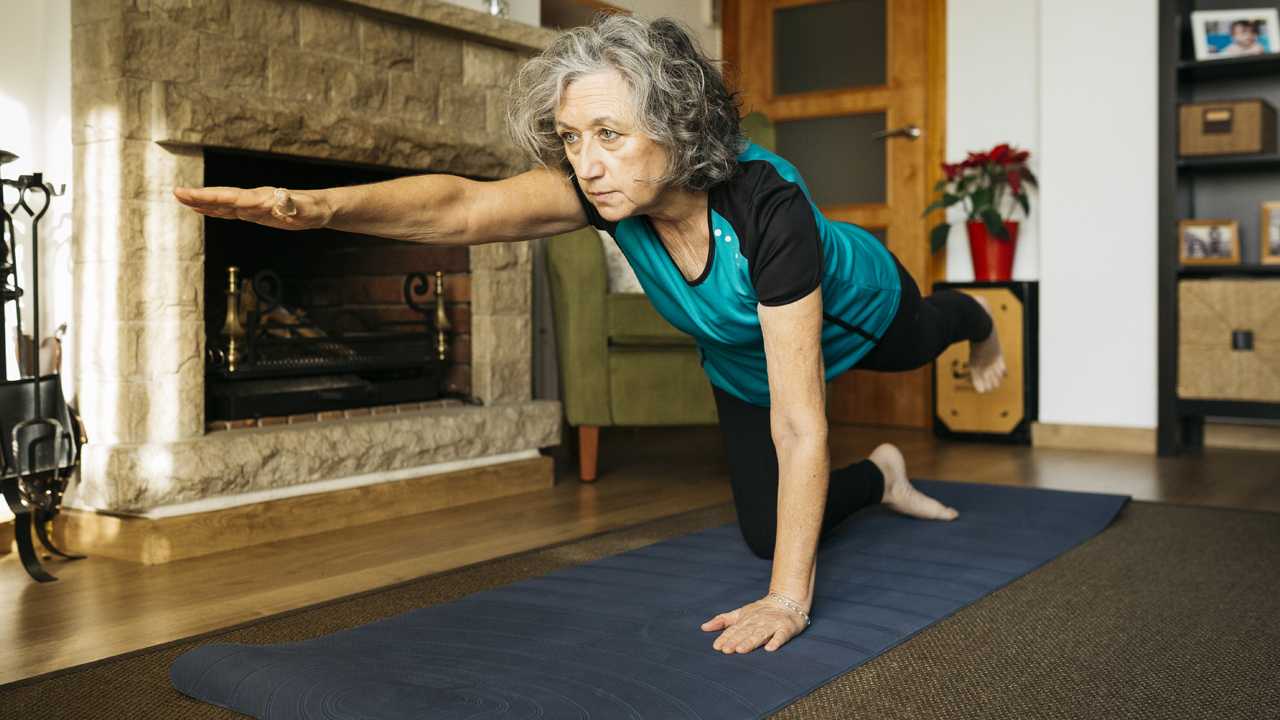 Person with short, curly gray hair in a balance pose on one knee and one hand on a yoga mat at home. 