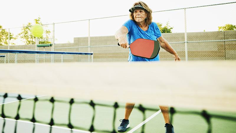 Older adult with a blue shirt and visor hitting a backhand shot during a pickleball game. 