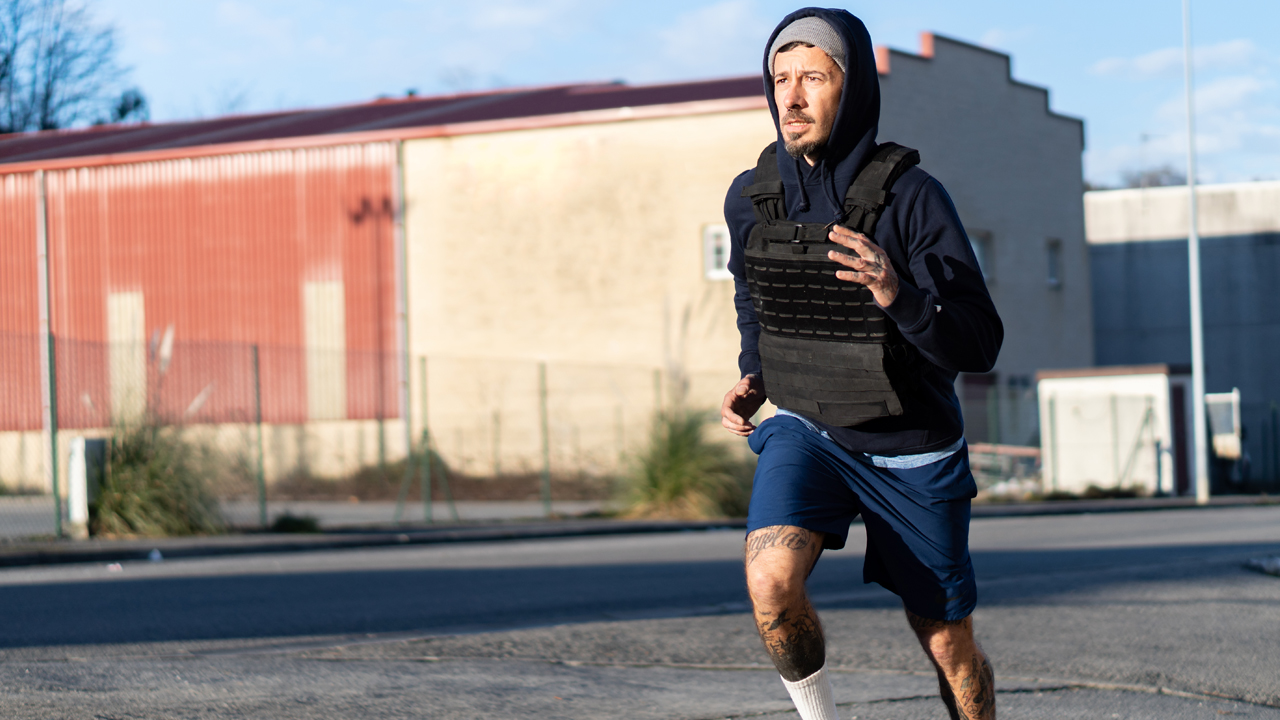 A person wearing a black weighted vest while running outside in shorts.