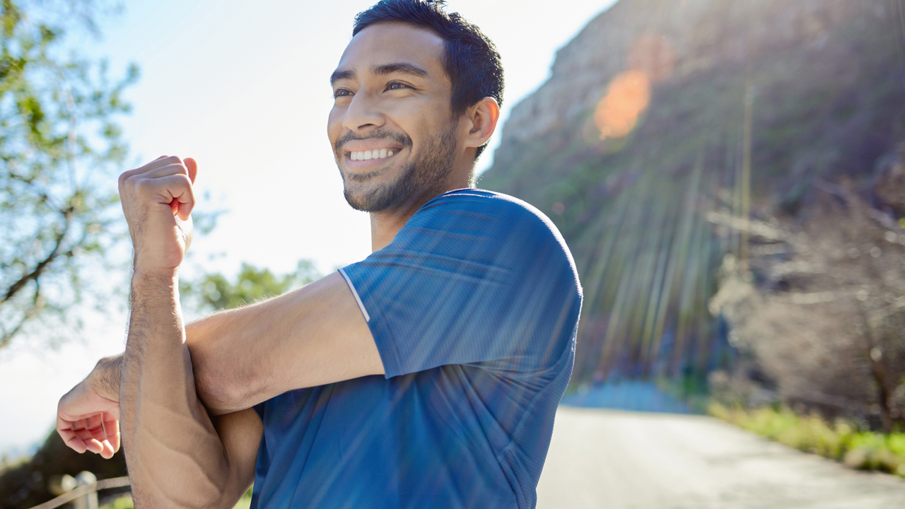 Person pictures from the mid-section up, outside in the sun, smiling, and using their right arm to pull their left across their chest and stretch.