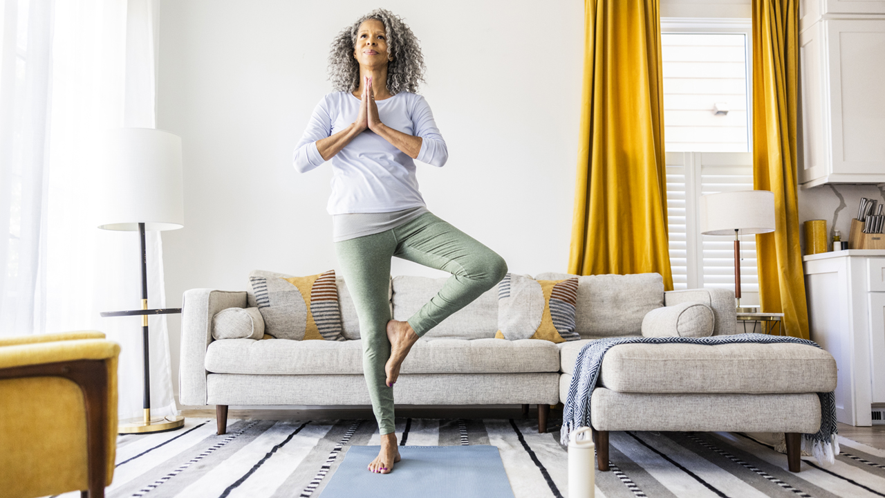 A person with long gray curly hair practices Tree Pose on a yoga mat in a living room. 