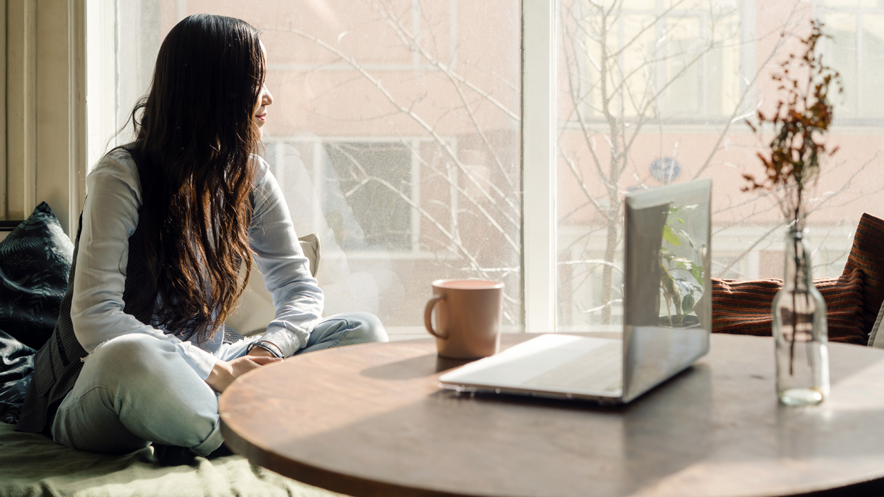 Person with long dark hair sits on a couch and looks out the window. A circular desk with a laptop, mug and plant are nearby