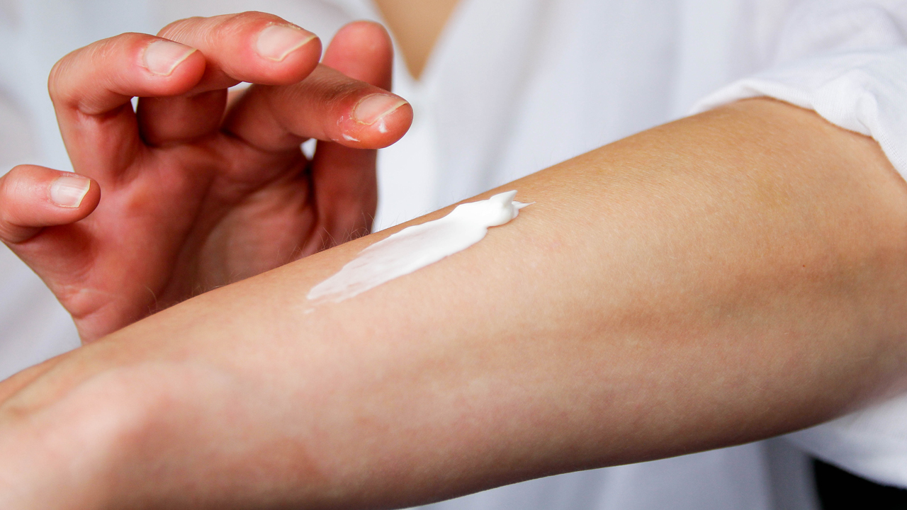 A closeup shot of a person's forearm with their hand, which looks somewhat red and irritated, spreading white lotion on it.