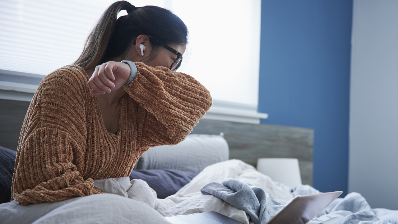 A person with long brown hair wearing glasses and an orange sweater coughs into sleeve while sitting on a bed. 