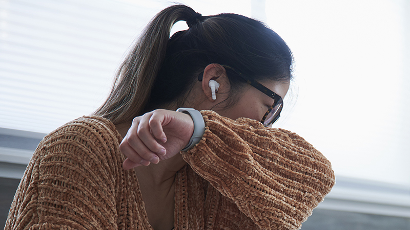 A person with long brown hair wearing glasses and an orange sweater coughs into sleeve while sitting on a bed. 