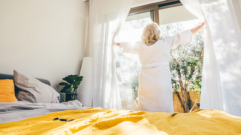 A person in a bedroom opens white curtains on a sunny morning. A bed with a yellow bedspread is nearby.