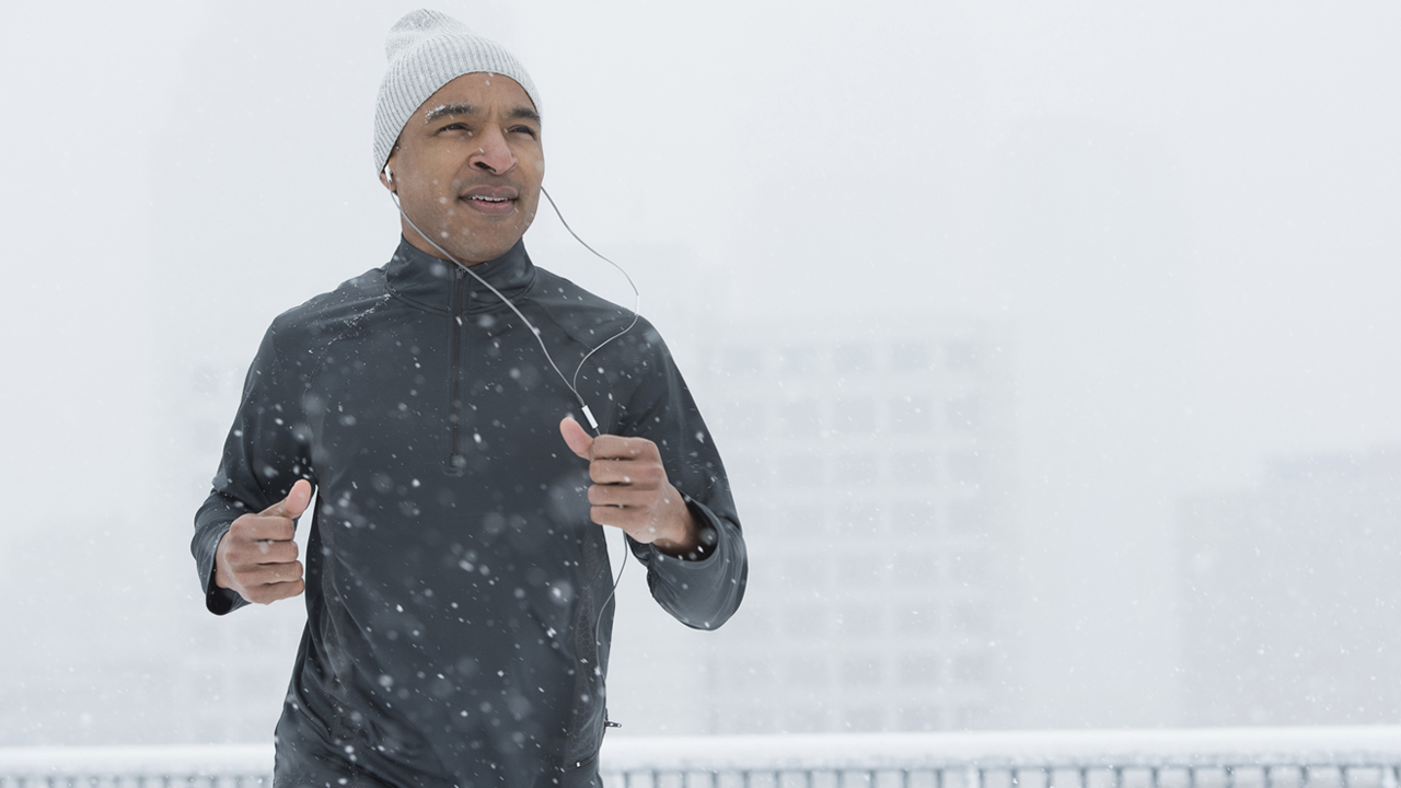 Torso length view of man running on snowy day wearing a white cap, dark gray jacket and wired ear buds. Faint office buildings in the background.