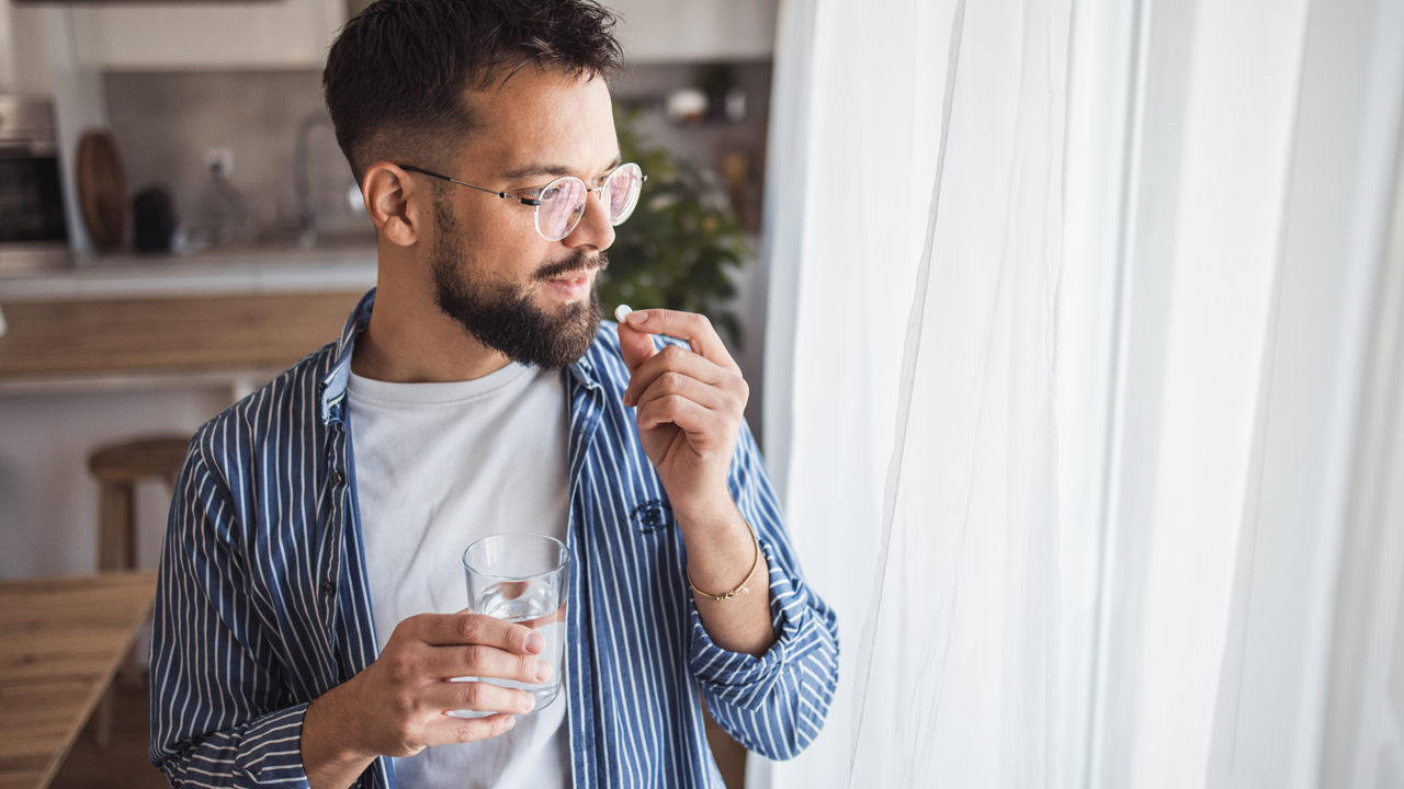 A person stands getting ready to take medication while holding a glass of water and a pill.