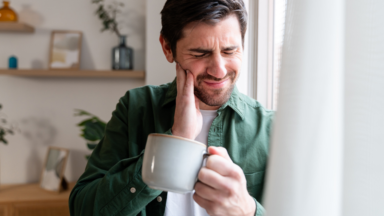 A person holding a cup of coffee and feeling their cheek with their hand.