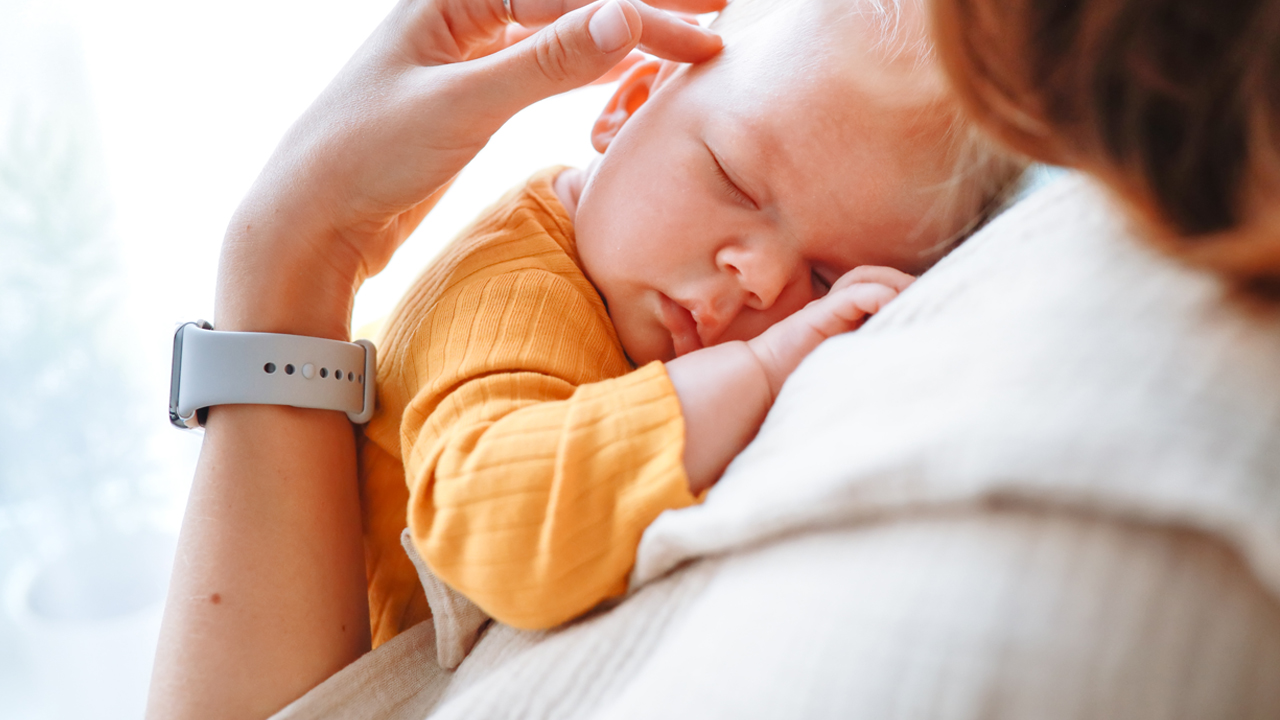 Adult with long brown hair wearing a white sweater and flexing their wrist, illustrating symptoms of mommy’s thumb or carpal tunnel syndrome, while holding a baby wearing a yellow outfit