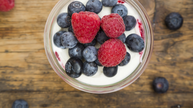 Glass of yogurt with fresh blueberries and raspberries on a wooden board.
