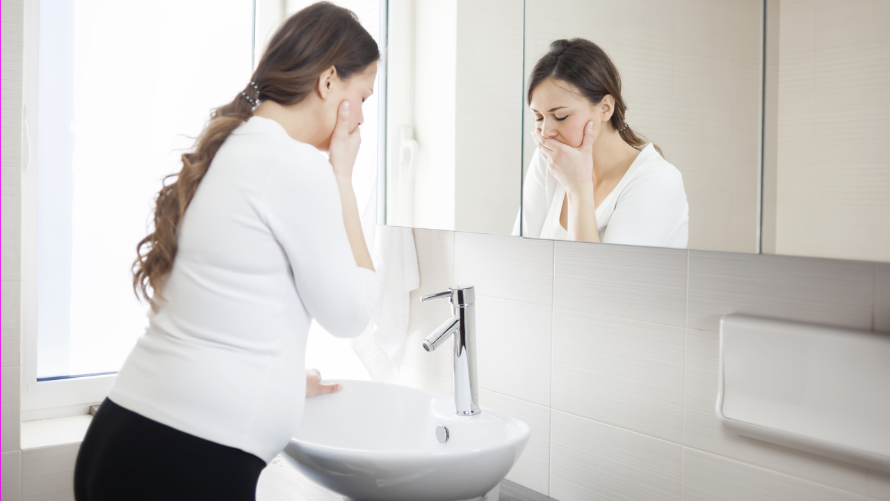 Person who is pregnant with long hair wearing a white shirt stands over a sink in a bathroom with one hand covering their mouth, indicating morning sickness and nausea during pregnancy