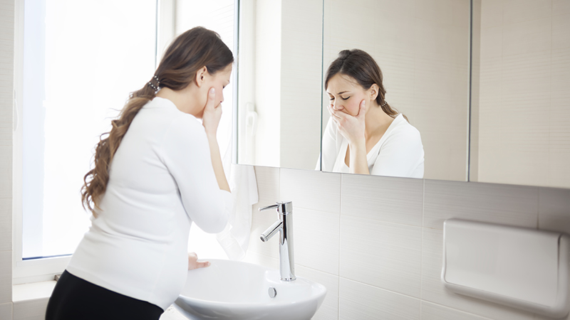 Person who is pregnant with long hair wearing a white shirt stands over a sink in a bathroom with one hand covering their mouth, indicating morning sickness and nausea during pregnancy