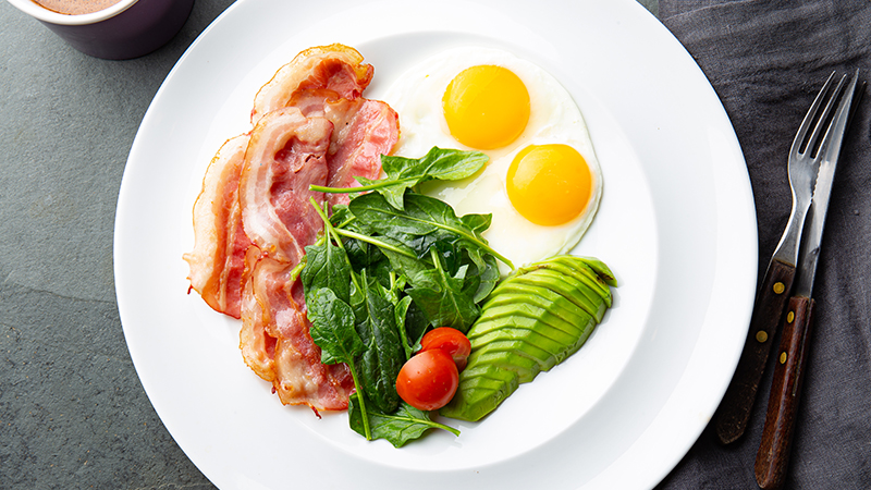 Overhead view of a white plate with sliced avocado, spinach, two grape tomatoes, three slices of bacon, and two sunny side up eggs.