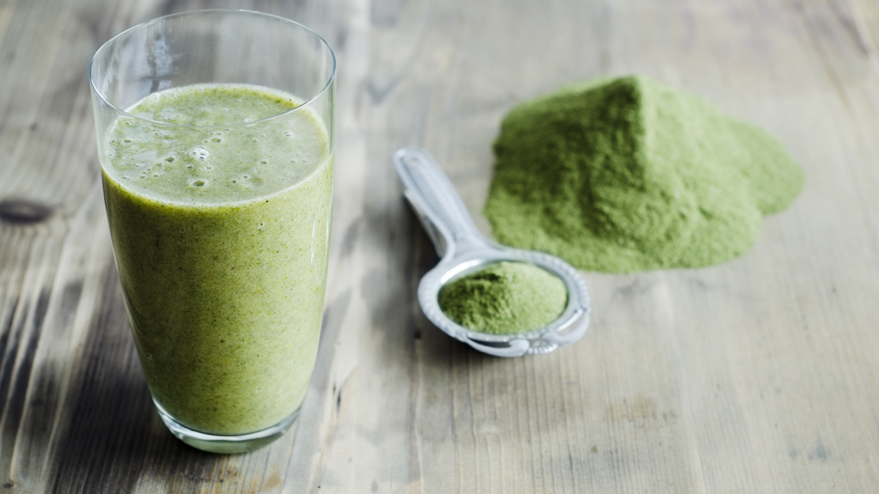 Green smoothie in a glass with green powder and a stainless steel spoon next to it on a wooden table. 