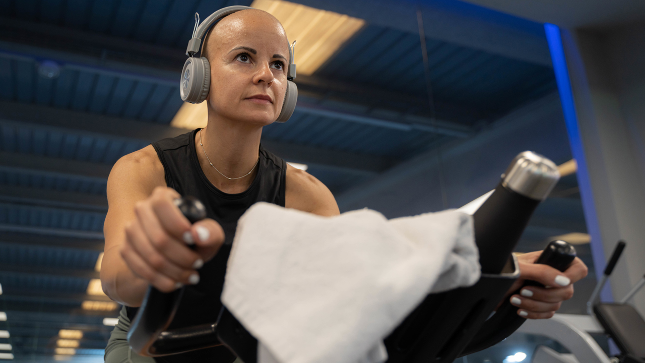 Woman without hair wearing headphones and concentrating while riding a stationary bike in a gym.