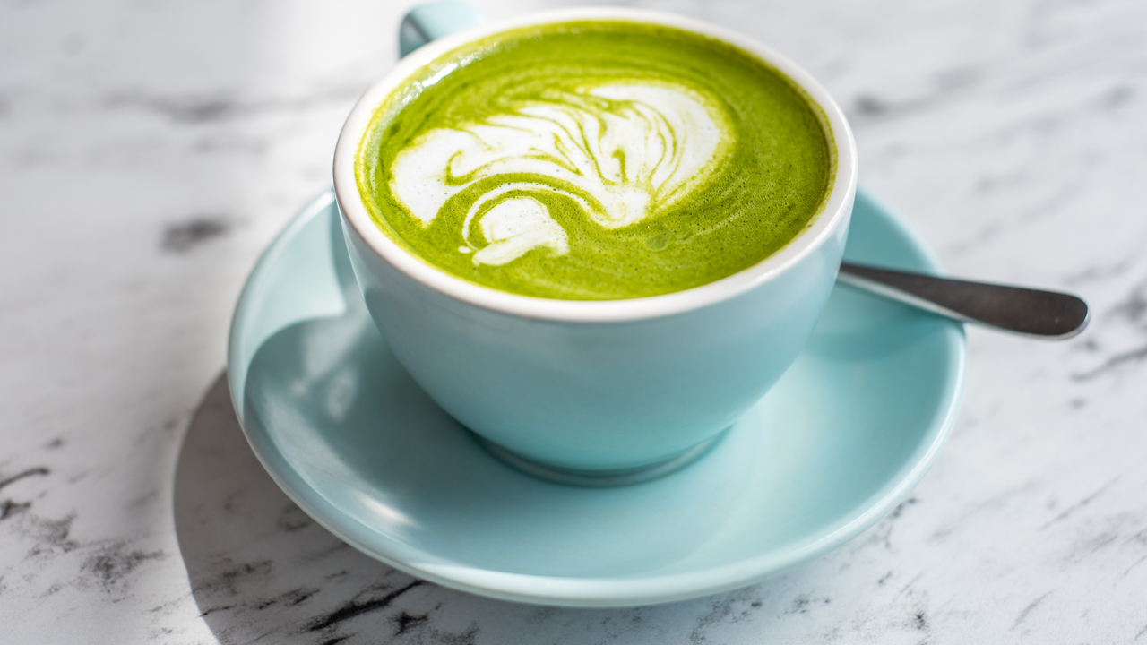 Green matcha latte in a blue mug on a plate with a spoon on a white and black quartz countertop. 