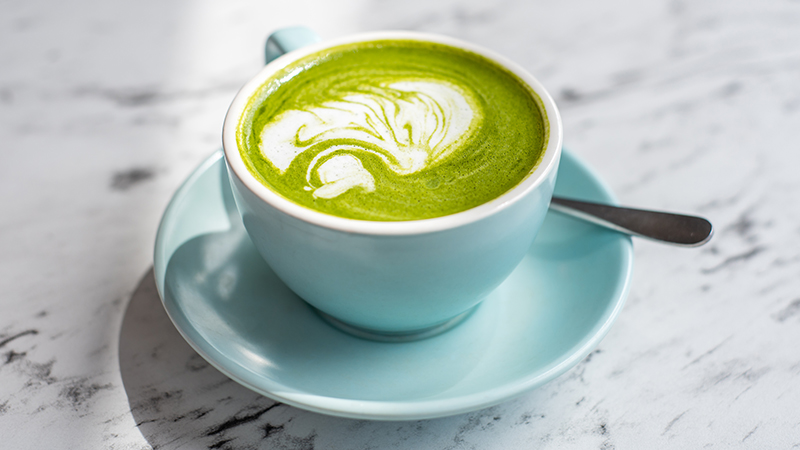 Green matcha latte in a blue mug on a plate with a spoon on a white and black quartz countertop. 