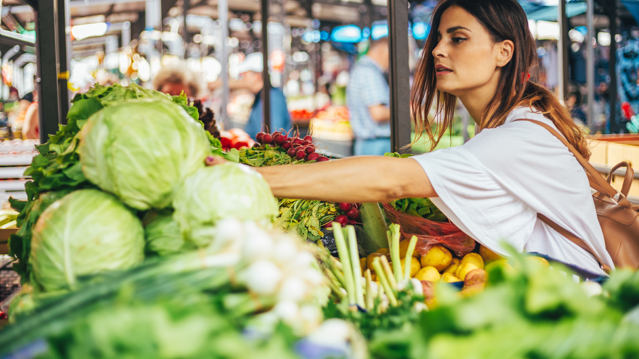 Person reaching for a head of cabbage in a grocery store. Other vegetables such as red radishes can be seen on the shelf as well.