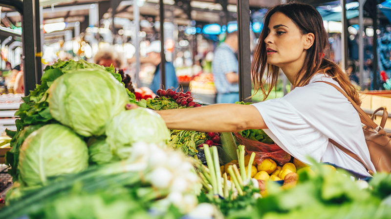 Person reaching for a head of cabbage in a grocery store. Other vegetables such as red radishes can be seen on the shelf as well.
